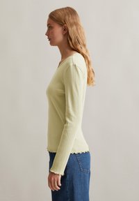 Lichtgroene geribbelde lange mouwtop met een v-hals en golvende zoom, gecombineerd met een donkerblauwe denimrok. De top heeft een aansluitende pasvorm.
