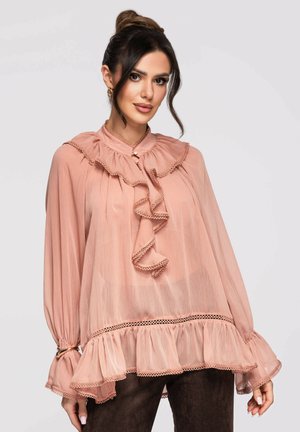 Mujer con cabello oscuro en un moño alto, vistiendo una blusa transparente rosa empolvado con volantes, mangas largas y detalles de botones de perla.