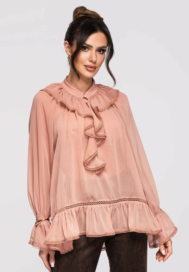 Femme aux cheveux foncés en chignon haut portant un chemisier transparent rose poudré à volants avec manches longues et détail de boutons en perle.