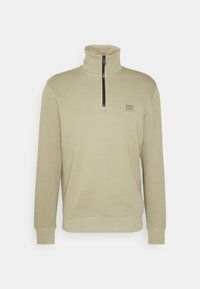 Béžový fleecový pulovr s vysokým límcem, polovičním zipem a čtvercovou nášivkou s logem na hrudi. Má žebrované manžety a lem. Hladká textura.