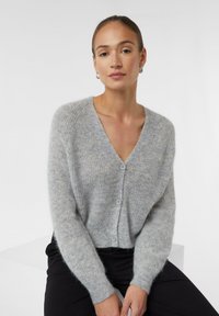 Lichtgrijze, fluffy cardigan met een V-hals, lange mouwen en drie knopen aan de voorkant. Zachte textuur met een relaxte pasvorm, gecombineerd met zwarte broek.
