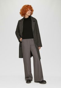 Grijze wollen jas met een geknipte kraag, gecombineerd met een zwarte coltrui en grijze wijde broek. Zwarte laarzen met rubberen zool maken de outfit compleet.