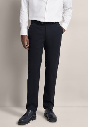 Mann trägt schwarze Stoffhose, schwarze Lederschuhe und ein weißes Hemd mit Knöpfen, steht mit einer Hand in der Tasche vor einem einfarbigen Hintergrund.