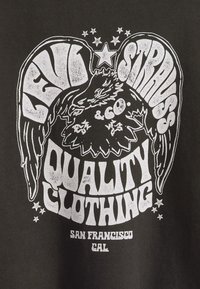 Fekete póló fehér grafikai dizájnnal, szárnyas motívummal, vastag betűkkel és csillagokkal. A felirat: "Levi Strauss & Co. Minőségi Ruházat."