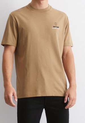 Man wearing a tan short-sleeve T-shirt with a small globe graphic and "mood worldwide" text on the left chest, paired with black pants.