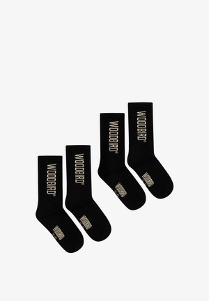 Vier schwarze Crew-Socken mit dem weißen Schriftzug "WOODBIRD", der vertikal an der Wade und horizontal nahe den Zehen gedruckt ist, auf weißem Hintergrund dargestellt.