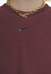 Bourgognefärgad fleece-tröja med rund halsringning och texturerad yta, med en subtil svart Nike-logga och bärs med ett guldhalsband.
