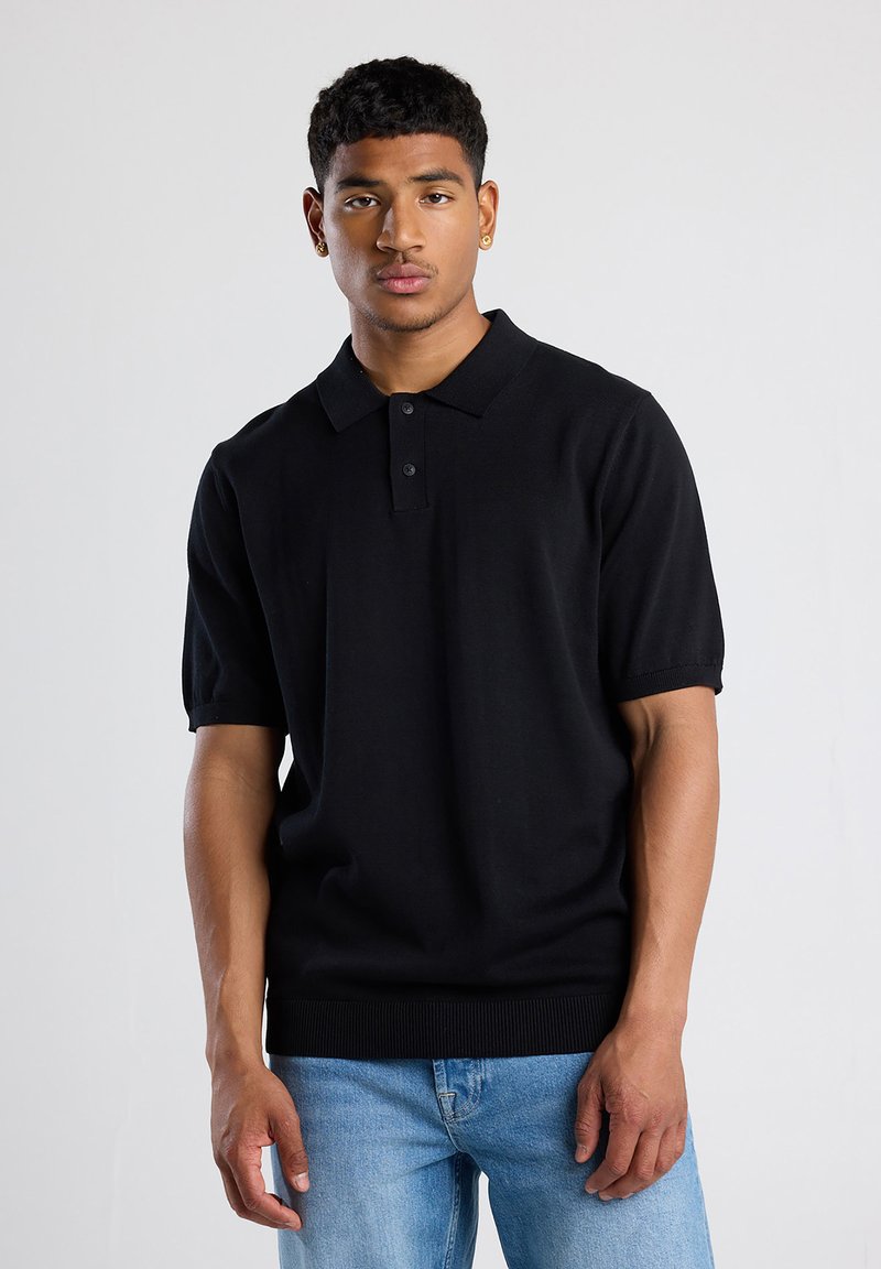 Black short-sleeve polo shirt made of knit fabric, featuring a buttoned placket and ribbed hem. Paired with light blue denim jeans.