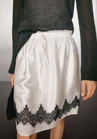 Weißer Satinrock mit schwarzer Spitzenborte am Saum, mit einem gerafften Bund und Kordelzug. Getragen mit einem dunklen Strickpullover.