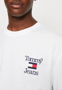 Camiseta blanca de algodón con el logotipo bordado de "Tommy Jeans" en azul marino, rojo y blanco. Cuello redondo y mangas cortas. Textura suave.