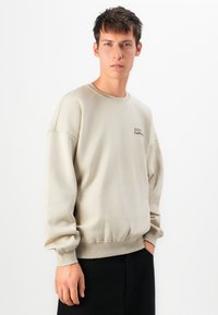 Jeune homme avec des cheveux bruns courts portant un sweat-shirt beige avec le texte "SOHO Coffee" et un pantalon noir, se tenant devant un fond blanc uni.