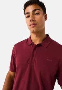 Camisa polo color burdeos hecha de una tela suave, con un cuello clásico, tres botones y el logo bordado "HUGO" en el lado izquierdo del pecho.