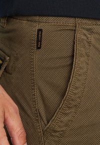 Bruine textuurbroek met een geweven patroon. Heeft een zijzak en een zwart label met "PANEL LEGEND" in gele stitch. Hand zichtbaar.