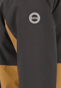 Giacca nera con una striscia diagonale gialla e un logo circolare grigio etichettato "ZIGZAG" sulla manica. Tessuto liscio e resistente.