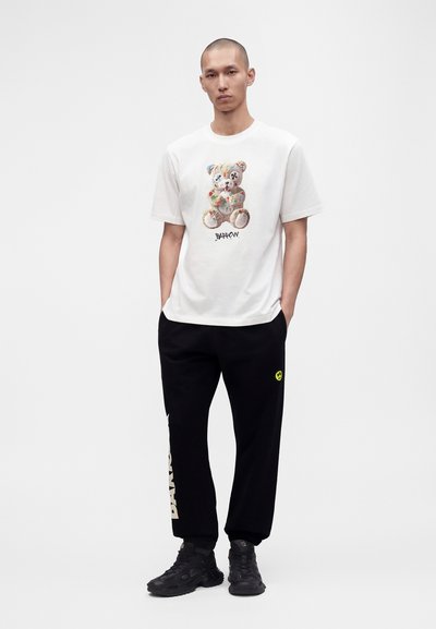 Homme debout, les mains dans les poches, portant un t-shirt blanc avec un imprimé ourson et un pantalon de survêtement noir avec un smiley jaune et des graphismes texte blancs.