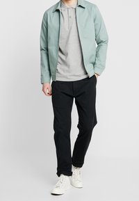 Světle zelená bomber bunda přes šedé polo tričko, doplněná černými kalhotami a bílými teniskami. Hladká tkanina a neformální design.