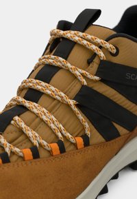 Nærbilde av brun tursko med oransje og beige mønstrede snørebånd, svarte stofføyler og teksturert grå såle synlig.