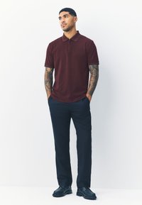 Polo bordeaux con un motivo leggermente strutturato, maniche corte e collo con zip, abbinato a pantaloni blu navy e scarpe nere.