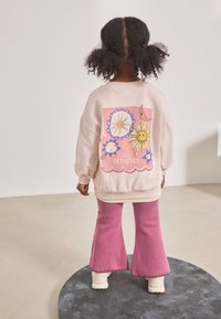 Ljusrosa sweatshirt med färggrafik av himlakroppar på ryggen, ihop med rosa utsvängda byxor och cremefärgade stövlar. Mjuk textur.