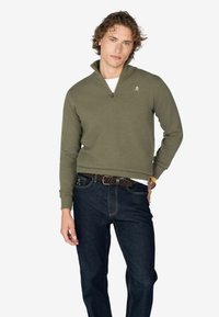 Sudadera de punto verde oliva con cremallera de un cuarto, puños de canalé y un pequeño logo en el pecho, combinada con pantalones de mezclilla oscuros.