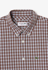 Camisa a cuadros marrón y azul claro, tipo button-up. Material de algodón con un corte regular, presenta un cuello, botones blancos y un logo de cocodrilo verde.