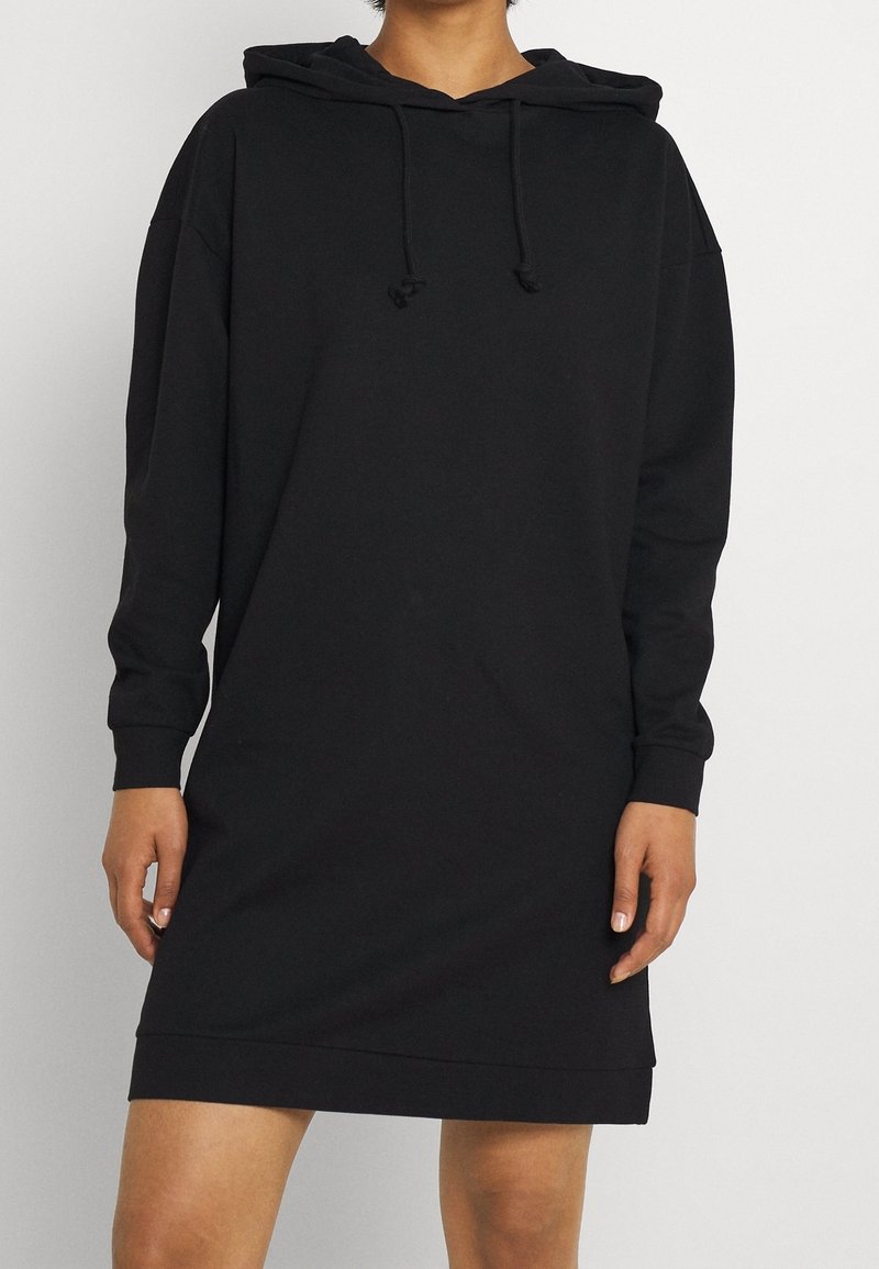 Personne portant une longue robe sweat à capuche noire avec manches longues et cordons, debout devant un fond clair uni.