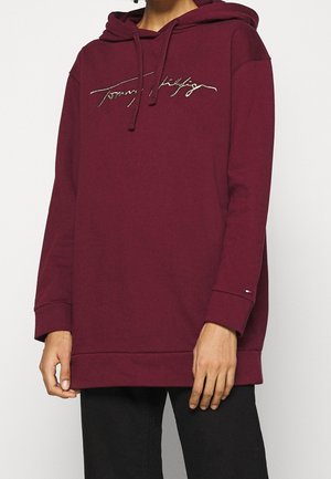 Personne portant un sweat à capuche bordeaux avec un cordon et un logo cursif sur la poitrine, associé à un pantalon noir sur un fond uni.