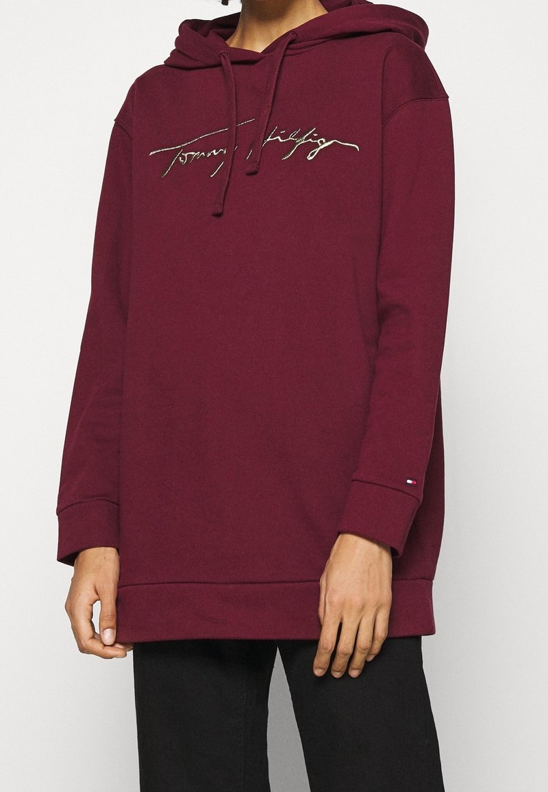 Personne portant un sweat à capuche bordeaux avec un cordon et un logo cursif sur la poitrine, associé à un pantalon noir sur un fond uni.
