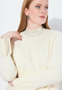 Sweter w kolorze kremowym z teksturowanym, wysokim kołnierzem, zdobionym wzorem małych prostokątnych akcentów. Gładki materiał, długie rękawy, luźny fason.