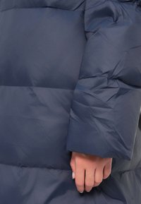 Tmavomodrý zateplený kabát zblízka ukazuje prešívanú textúru, hladký povrch a mierne voľný rukáv s rukou položenou na látke.