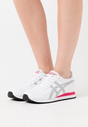 Person som bär vita och grå sneakers med rosa detaljer, stående på en vit bakgrund.