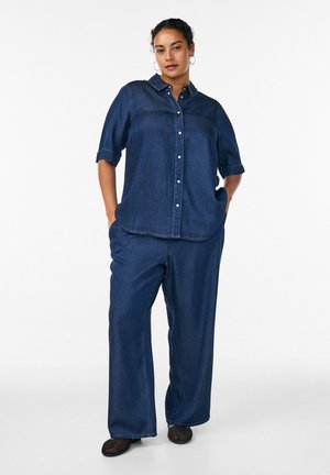 Žena stojí s rukami v vreckách, oblečená v tmavomodrej rifľovej košeli na gombíky a zodpovedajúcich širokých nohaviciach na bielom jednoliatom pozadí.