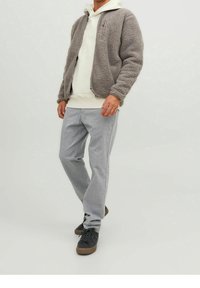 Uomo che indossa una giacca in pile grigia con zip, una felpa bianca con cappuccio, pantaloni grigio chiaro e sneaker nere in piedi contro uno sfondo bianco semplice.
