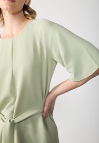Robe vert clair en tissu texturé, avec un col rond et des manches cloche. Détail ceinture nouée avec poches apparentes.
