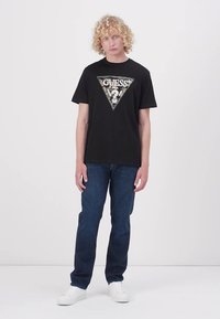 Svart bomulls T-skjorte med et trekantet grafisk design, med "GUESS" i metallisk skrift. Kombinert med blå denim jeans og hvite sneakers.