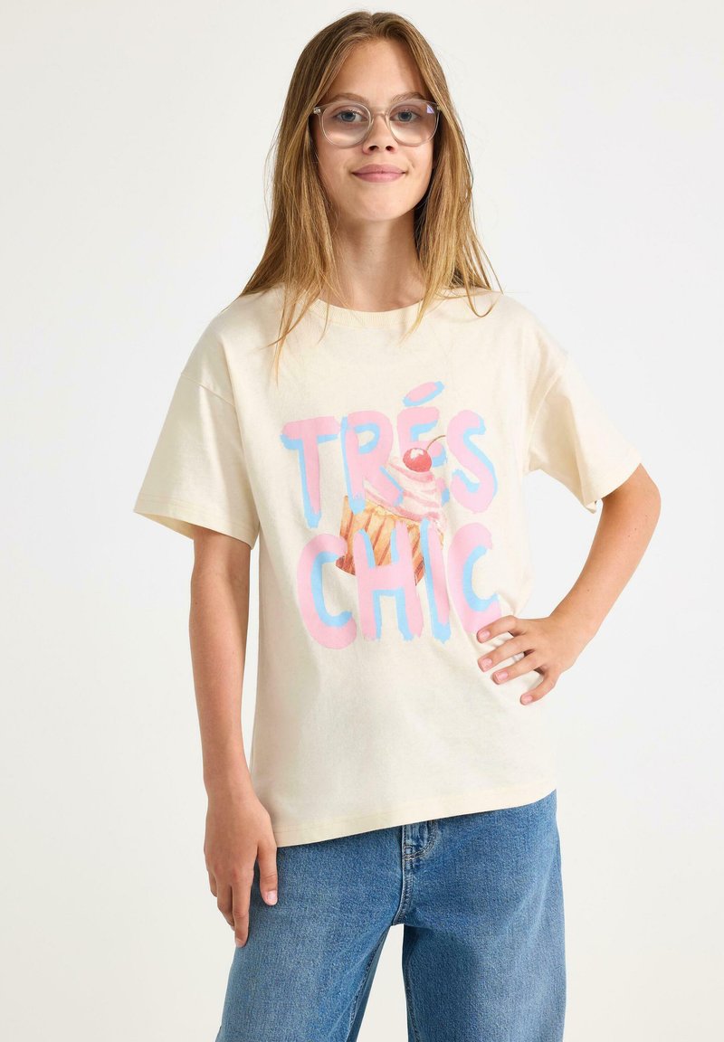 Krämfärgad bomull t-shirt med texten "TRÈS CHIC" i rosa och blå, tillsammans med en grafik av en glasskon. Kort ärm och avslappnad passform.
