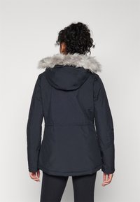 Parka negra con capucha forrada de pelo sintético, que presenta una cintura ajustada, mangas largas y una textura suave. La vista trasera muestra un diseño simple.