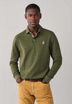 Joven con una camisa polo de manga larga color verde oliva con un logo bordado y pantalones amarillos mostaza, de pie con las manos en los bolsillos.