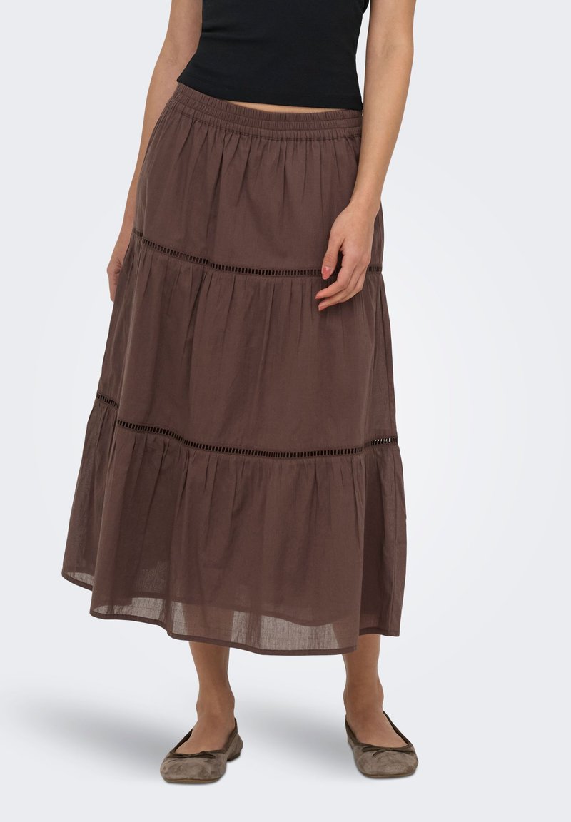 Femme portant une jupe midi marron à volants avec des inserts en dentelle, associée à des chaussures plates taupe et un haut noir sans manches, debout sur fond blanc.