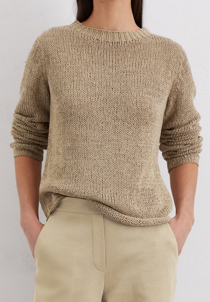 Femme portant un pull en maille beige aux manches retroussées et un pantalon beige, debout les mains dans les poches contre un mur blanc uni.