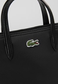 Sac à main noir texturé avec des poignées en cuir noir et un petit logo crocodile vert brodé près de la fermeture éclair supérieure.