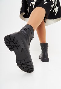Botas de tobillo de cuero negro con un acabado texturizado, punta redonda y suela resistente con profundos surcos. Incluye una correa decorativa en el tobillo.