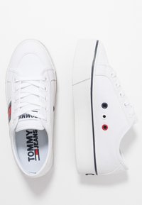 Sneakers bianche in tela con suola spessa. Presentano dettagli in rosso e blu navy, design con lacci e un logo sulla linguetta. Suola in gomma con finitura testurizzata.