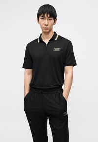 Μαύρο πόλο με φερμουάρ, γιακά με χρυσές λεπτομέρειες και λογότυπο EA7, συνδυασμένο με μαύρο υφασμάτινο παντελόνι jogger με πλαϊνές τσέπες.