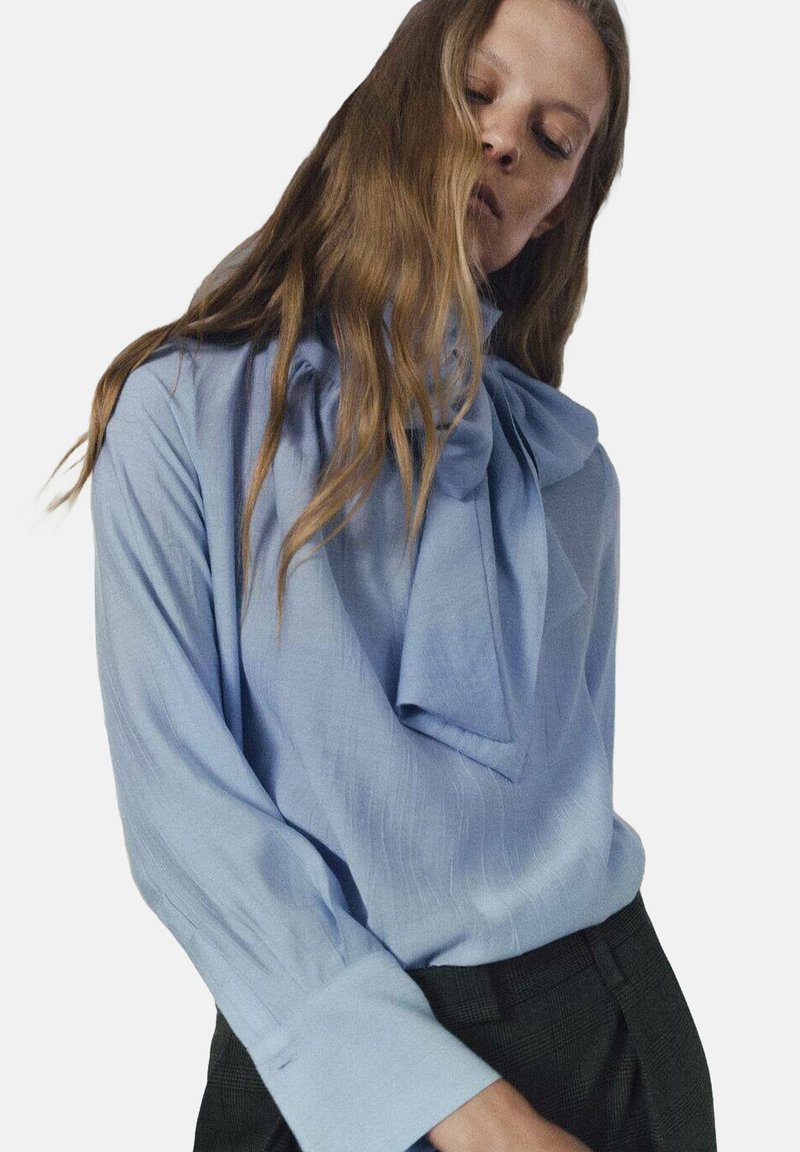 Blusa azul clara con un gran lazo en el escote, mangas largas y un tejido texturizado. Combinada con pantalones de cuadros oscuros.