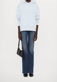 Osoba v světle modré mikině s kapucí, rozšiřujících se modrých džínách, černých botách, držící malou černou kabelku u své strany na jednoduchém pozadí.