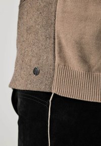 Primer plano de un jersey de punto beige con dobladillo acanalado y panel lateral texturizado sobre pantalones negros con detalle de costura blanca.