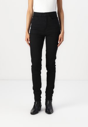 Femme portant un jean skinny noir taille haute et des bottines noires, debout devant un fond clair uni.