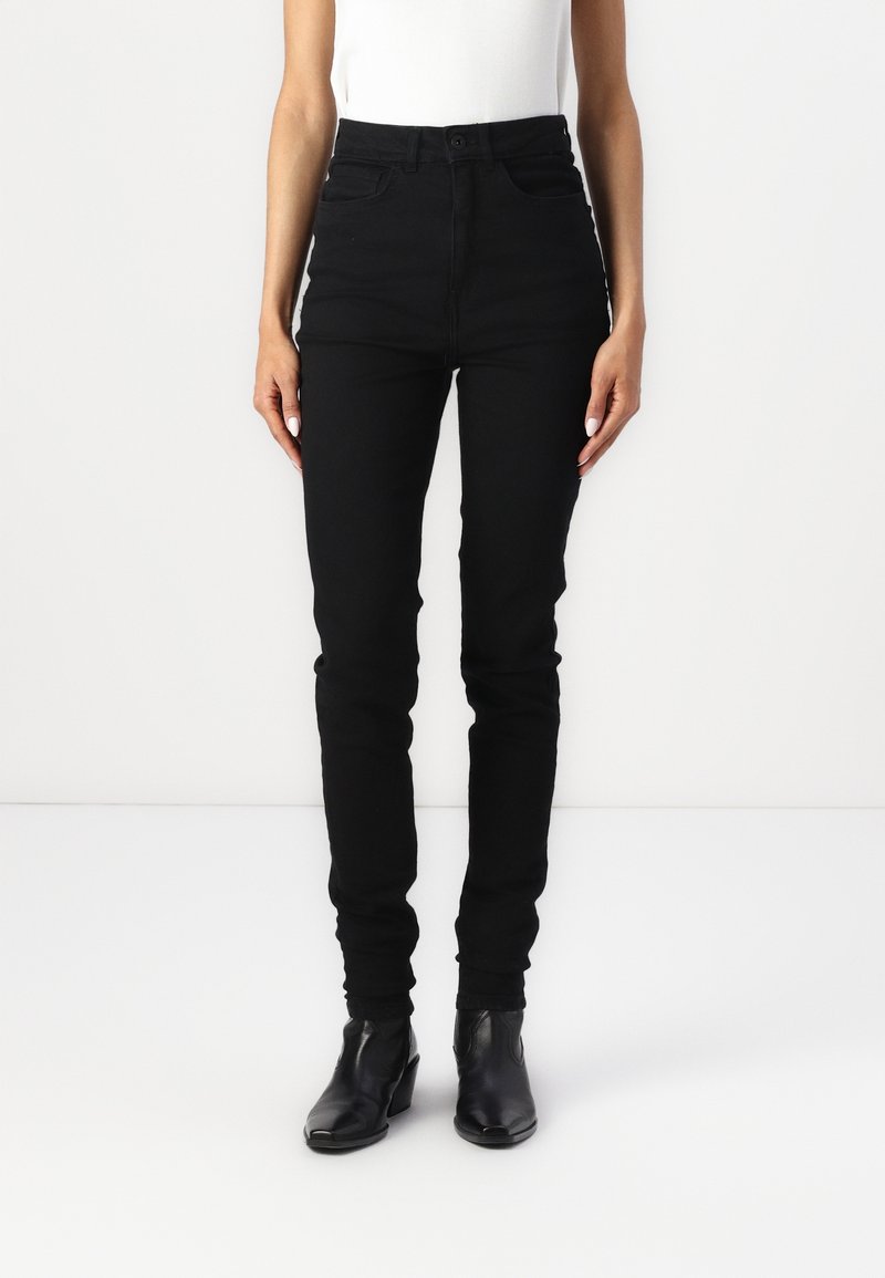 Femme portant un jean skinny noir taille haute et des bottines noires, debout devant un fond clair uni.