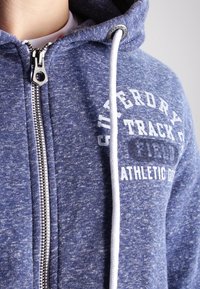 Marineblauer Kapuzenpullover mit Reißverschluss aus strukturiertem Stoff und weißen Kordeln, mit dem Aufdruck "SUPERDRY TRACK" auf der Brust.